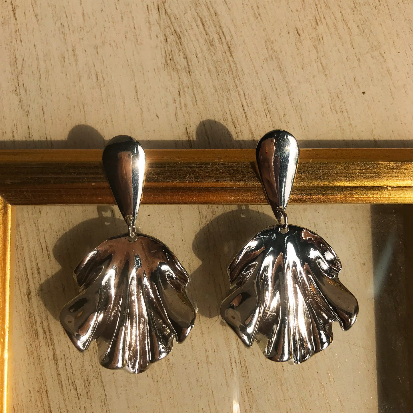 Two sculptural metal drop earrings, one silver and one gold, with a flowing, draped design, displayed against a framed background.
