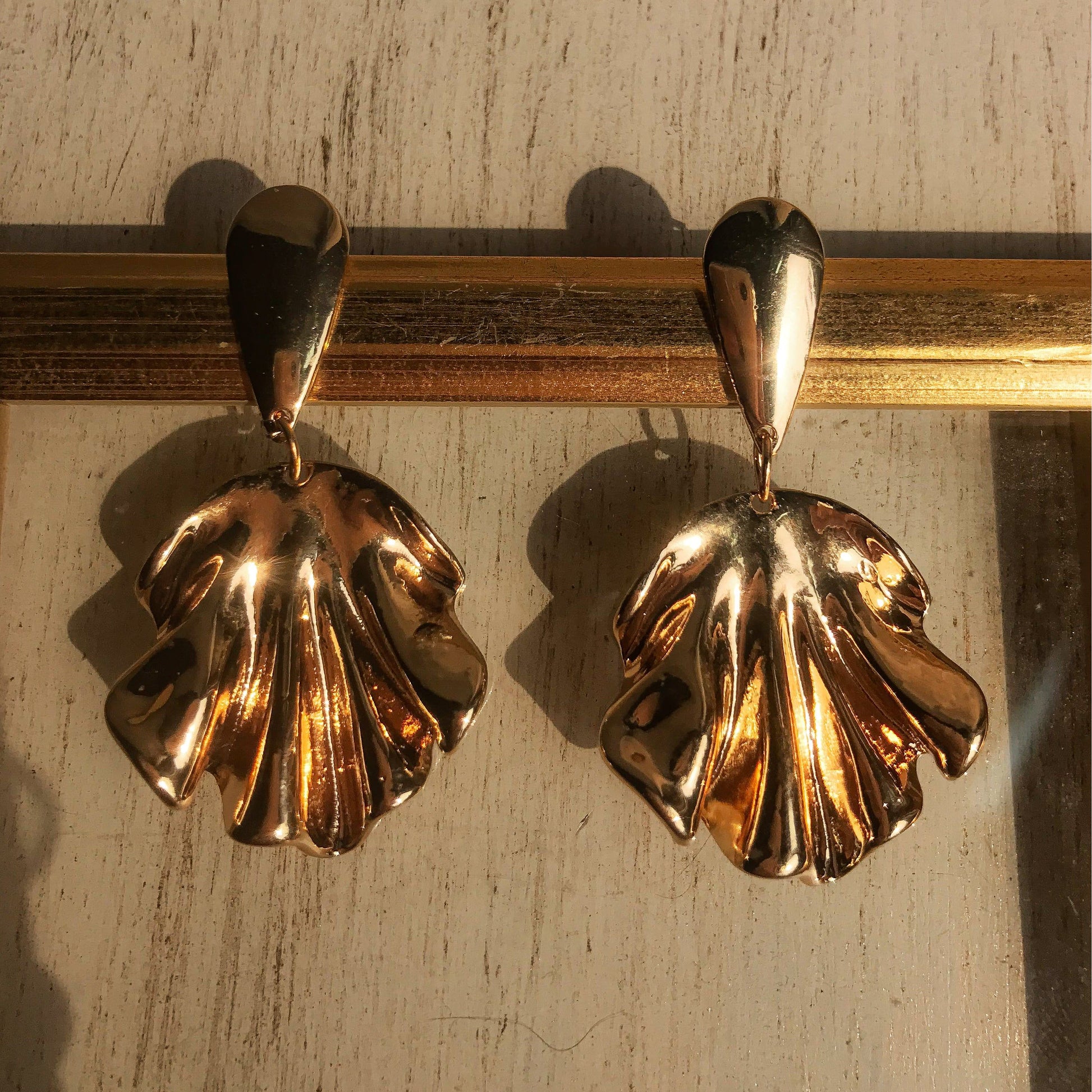 Two sculptural metal drop earrings, one silver and one gold, with a flowing, draped design, displayed against a framed background.