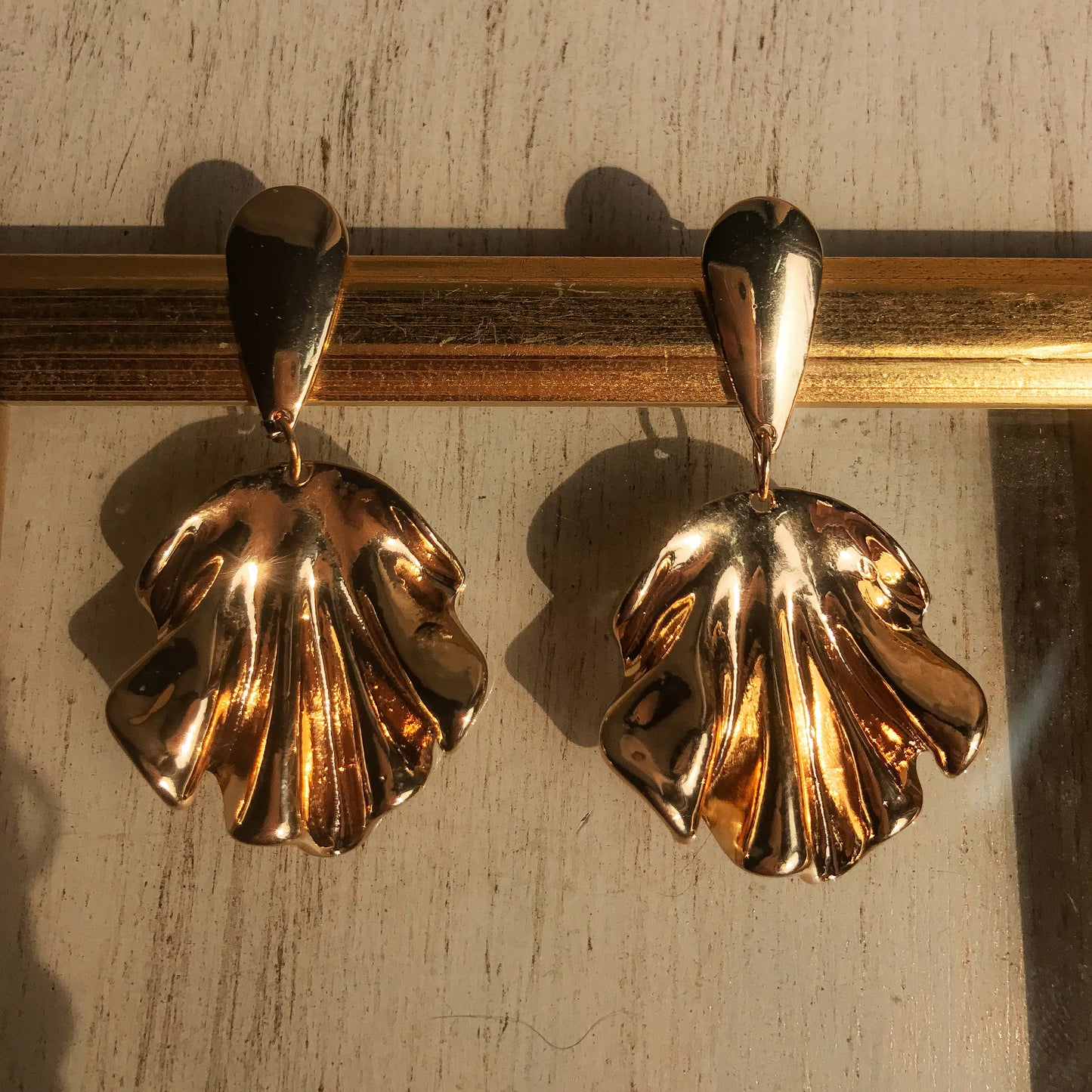 Two sculptural metal drop earrings, one silver and one gold, with a flowing, draped design, displayed against a framed background.