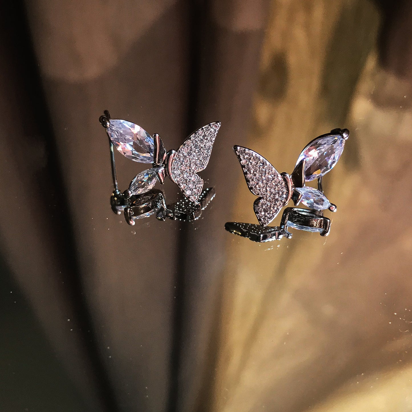 Two butterfly-shaped stud earrings, one silver and one gold, with marquise-cut stones forming the wing edges and smaller stones filling the inner wings, displayed on a reflective surface.