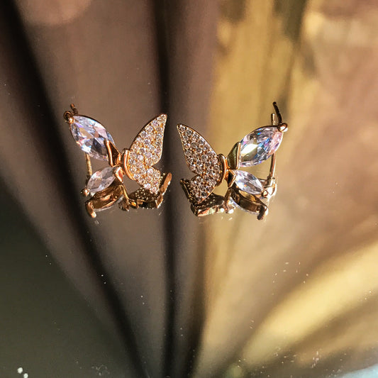 Two butterfly-shaped stud earrings, one silver and one gold, with marquise-cut stones forming the wing edges and smaller stones filling the inner wings, displayed on a reflective surface.