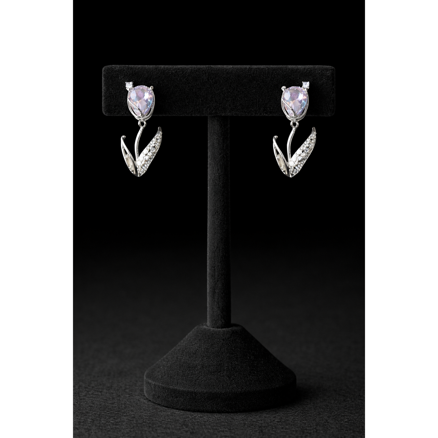 Silver tulip-shaped drop earrings with soft lavender crystal stones and pavé leaf detailing displayed on a black velvet T-bar stand against a dark black background in high-definition
