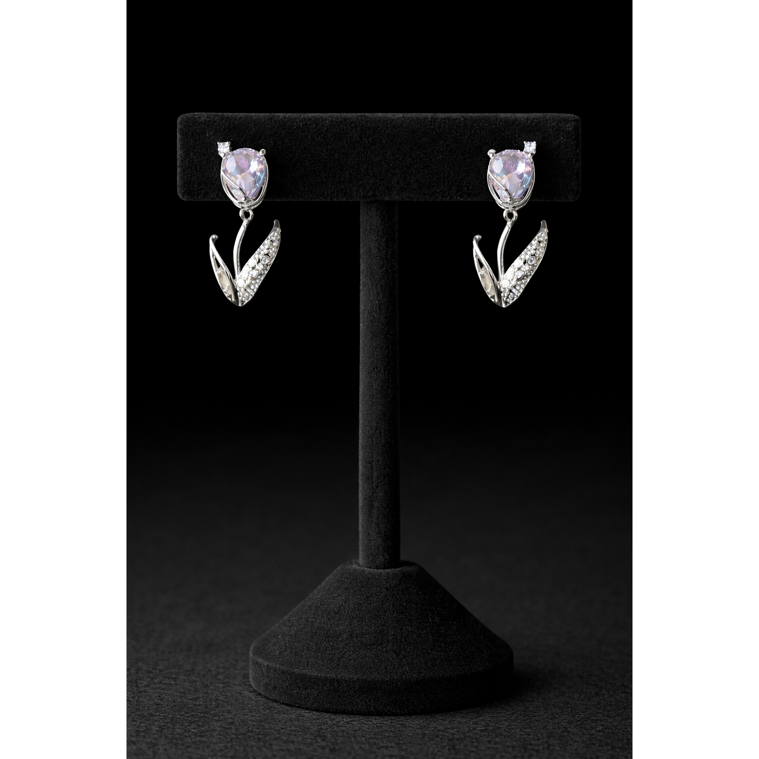 Silver tulip-shaped drop earrings with soft lavender crystal stones and pavé leaf detailing displayed on a black velvet T-bar stand against a dark black background in high-definition