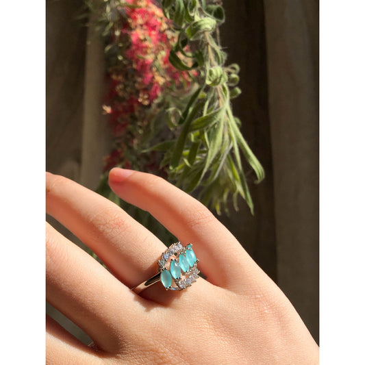 A close-up of a sparkling silver-tone ring featuring a large round clear stone surrounded by a halo of smaller clear stones, with more small clear stones set along the band.