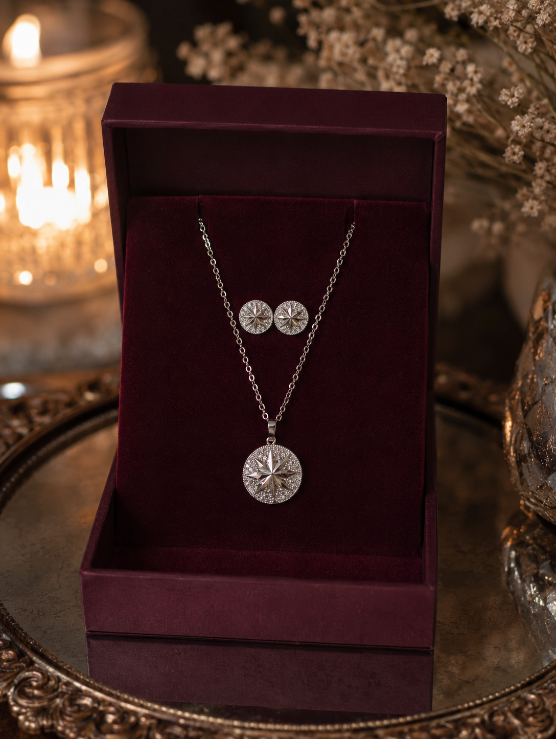 Silver starburst pendant and matching stud earring set displayed in a premium burgundy velvet jewelry box with warm candlelight and decorative dried flowers in the background.