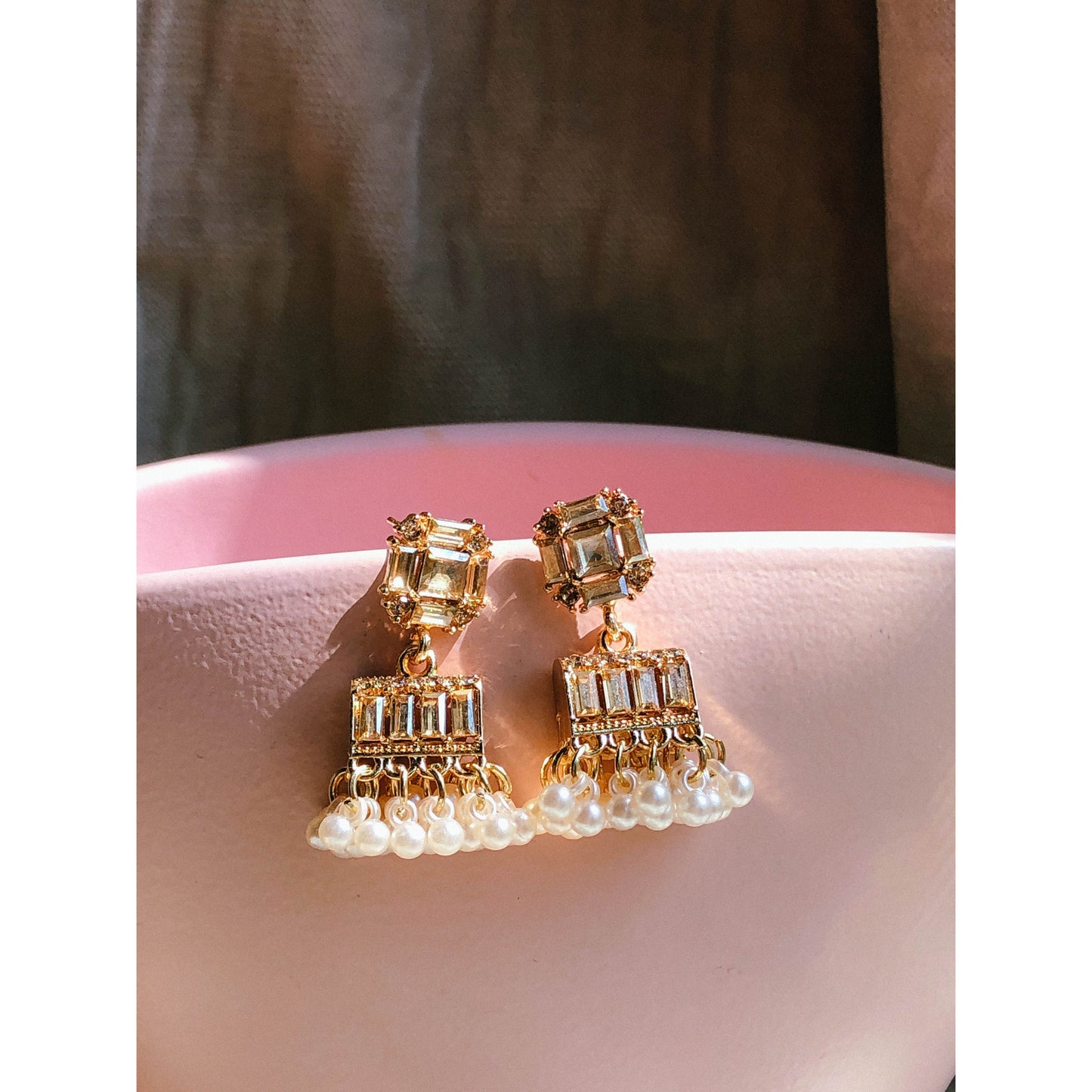 Gold-plated pearl jhumka earrings with crystal stones resting on a pink surface