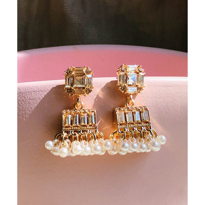 Gold-plated pearl jhumka earrings with crystal stones resting on a pink surface