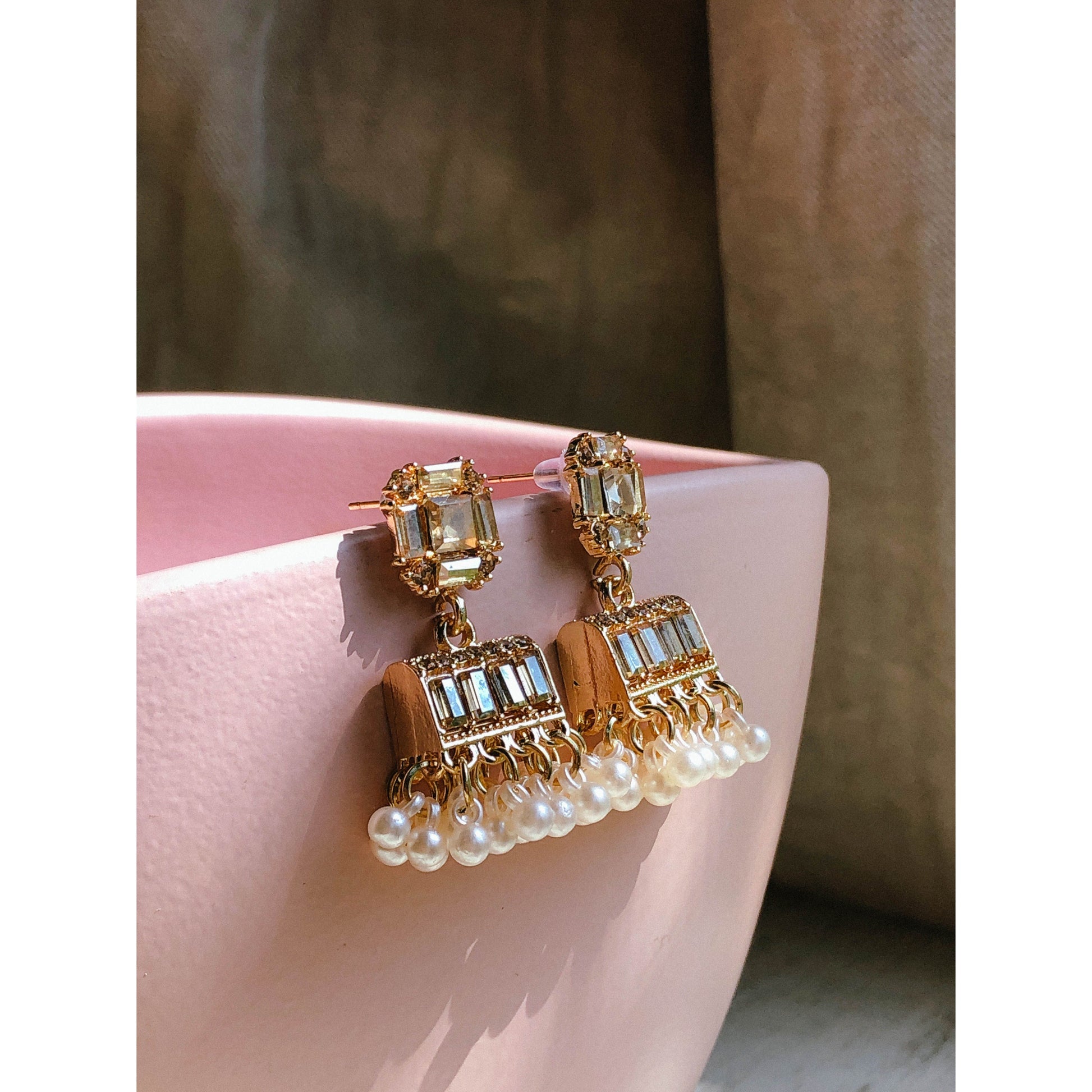 Gold-plated pearl jhumka earrings with crystal stones resting on a pink surface