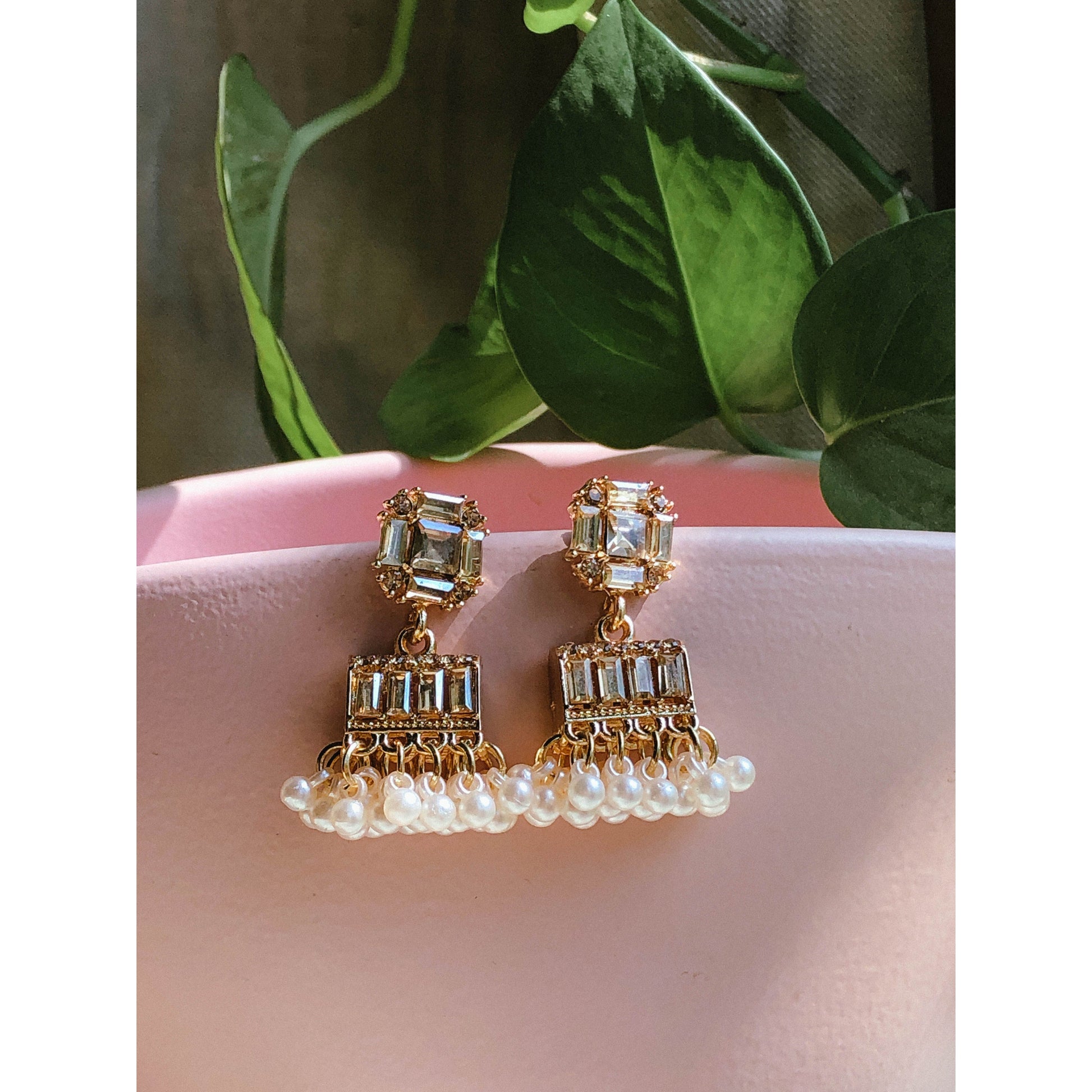 Gold-plated pearl jhumka earrings with crystal stones resting on a pink surface