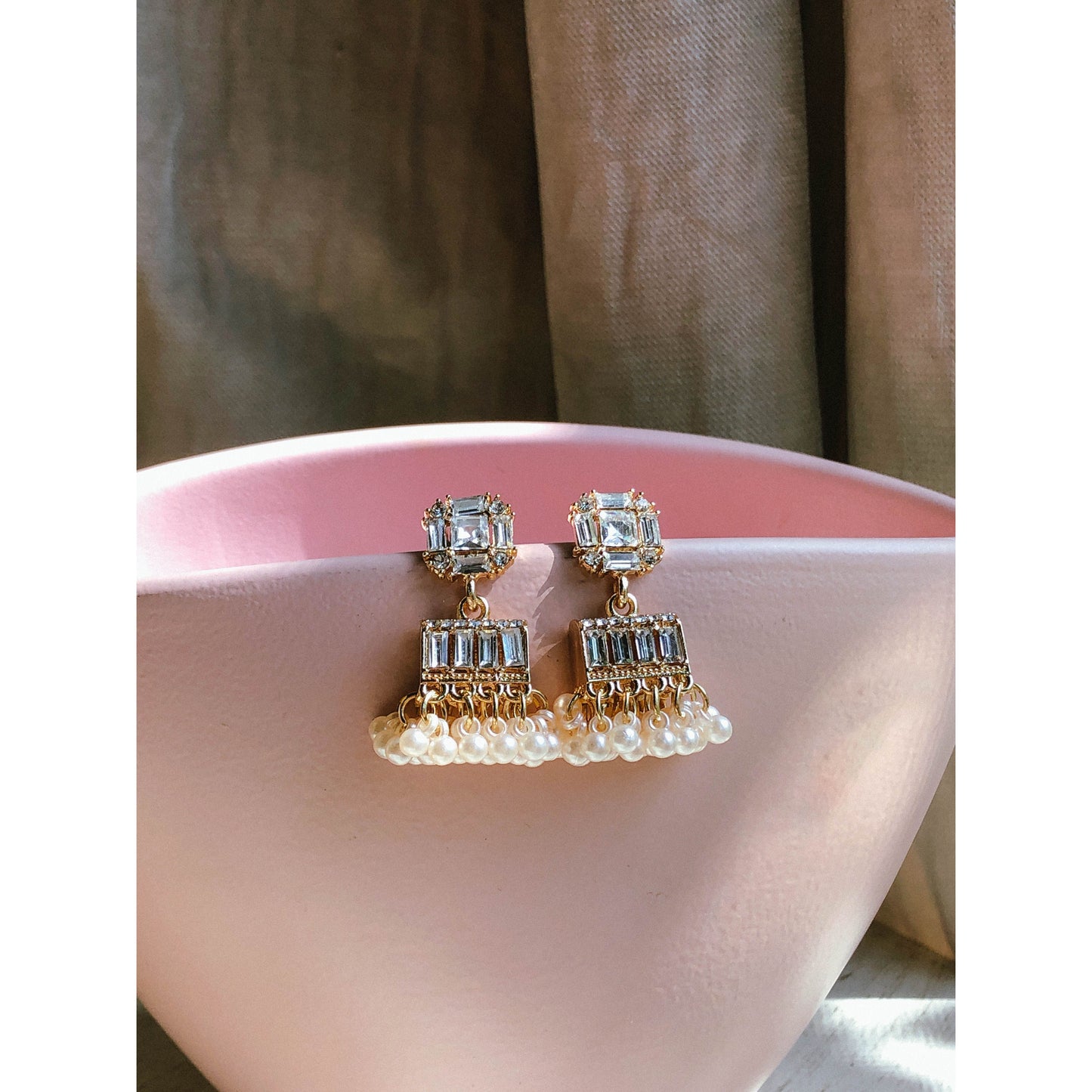 Gold-plated pearl jhumka earrings with crystal stones resting on a pink surface