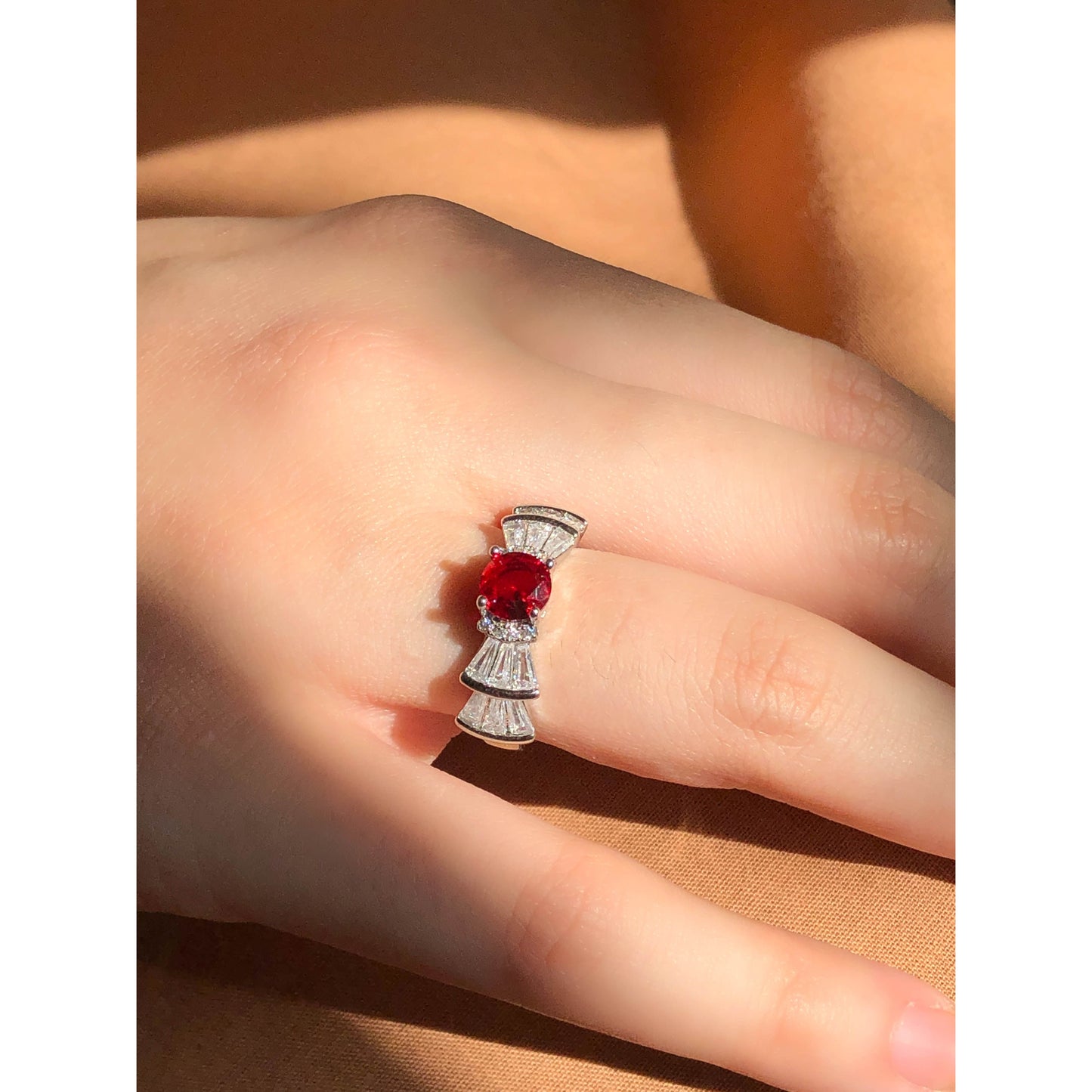 A silver-tone ring featuring an oval-cut gemstone in aqua and blush options, accented by stacked baguette-cut crystals on each side, worn on a hand with warm-toned fabric in the background.