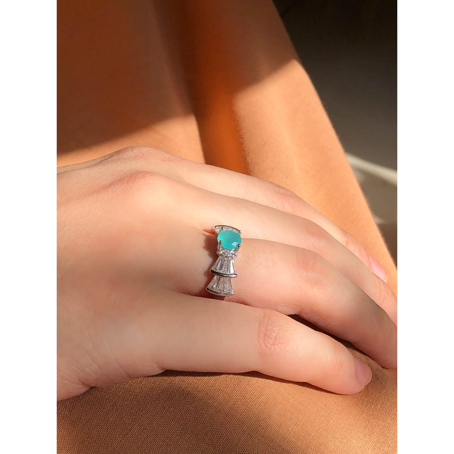 A silver-tone ring featuring an oval-cut gemstone in aqua and blush options, accented by stacked baguette-cut crystals on each side, worn on a hand with warm-toned fabric in the background.