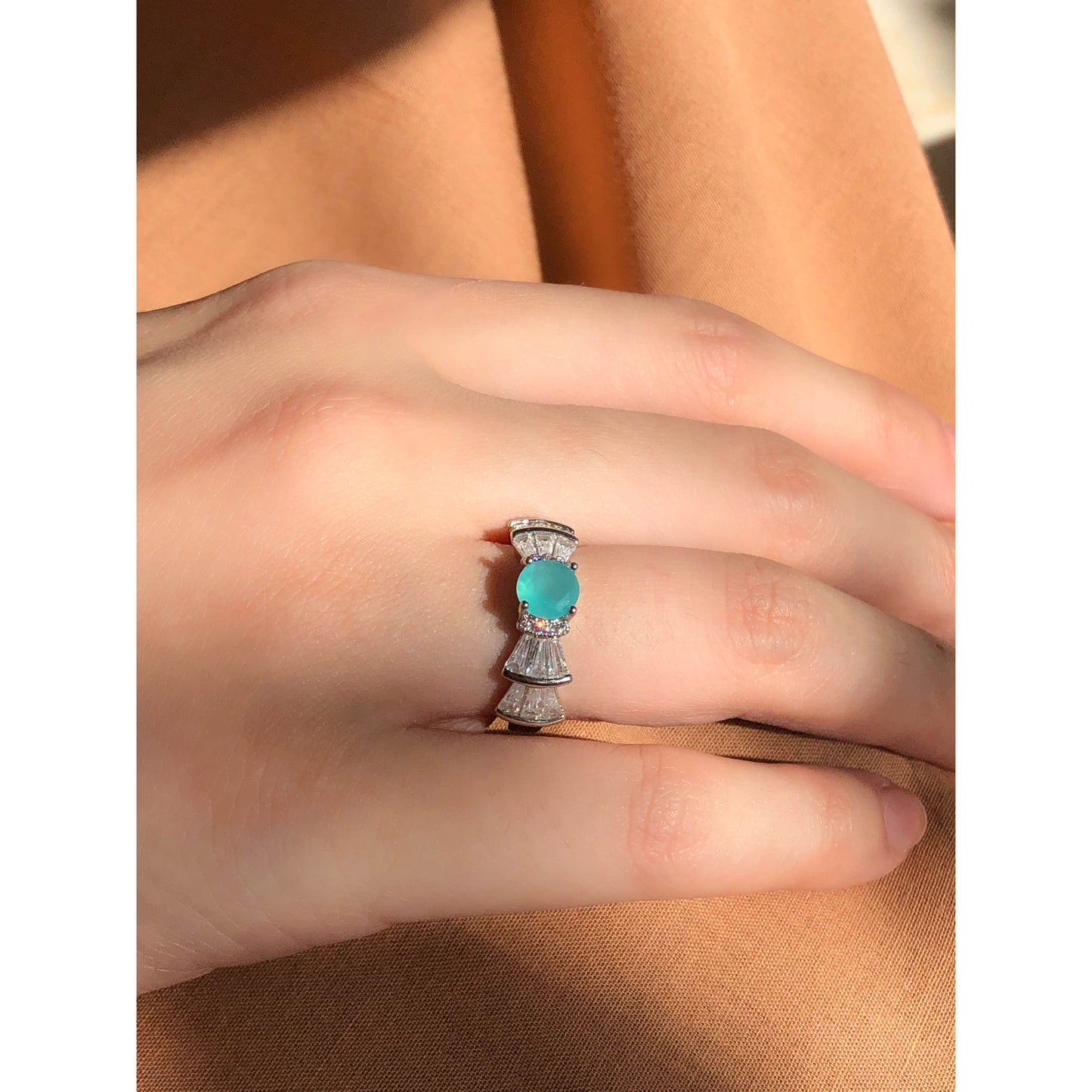 A silver-tone ring featuring an oval-cut gemstone in aqua and blush options, accented by stacked baguette-cut crystals on each side, worn on a hand with warm-toned fabric in the background.