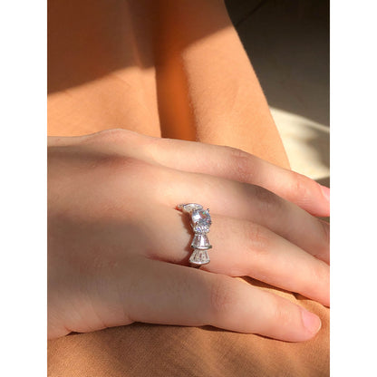 A silver-tone ring featuring an oval-cut gemstone in aqua and blush options, accented by stacked baguette-cut crystals on each side, worn on a hand with warm-toned fabric in the background.