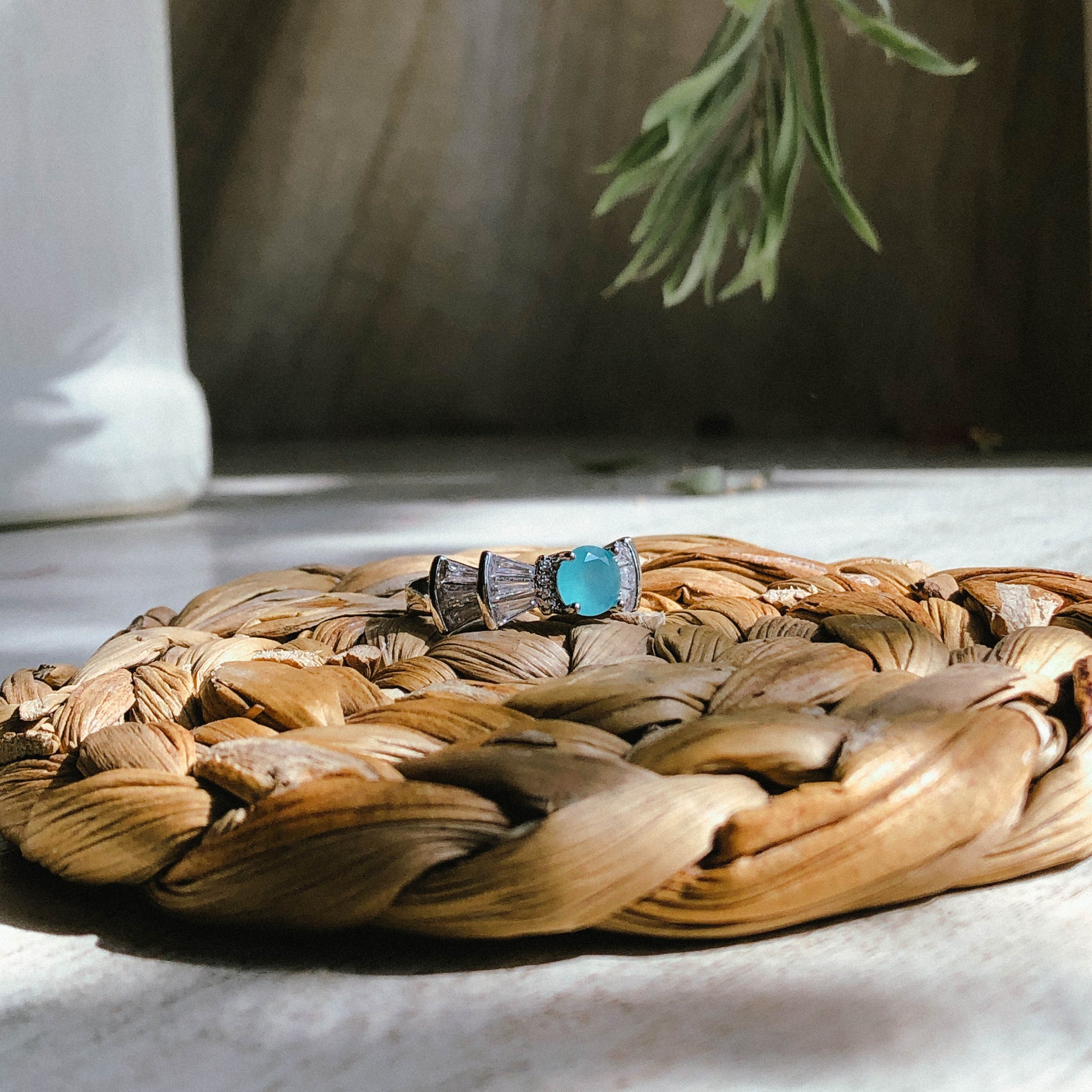 A silver-tone ring featuring an oval-cut gemstone in aqua and blush options, accented by stacked baguette-cut crystals on each side, worn on a hand with warm-toned fabric in the background.