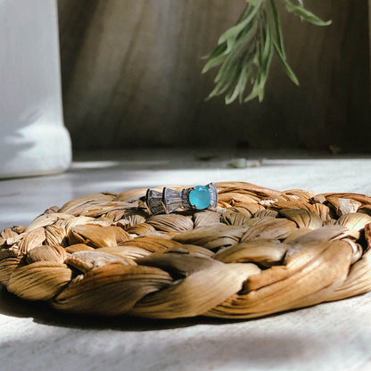 A silver-tone ring featuring an oval-cut gemstone in aqua and blush options, accented by stacked baguette-cut crystals on each side, worn on a hand with warm-toned fabric in the background.