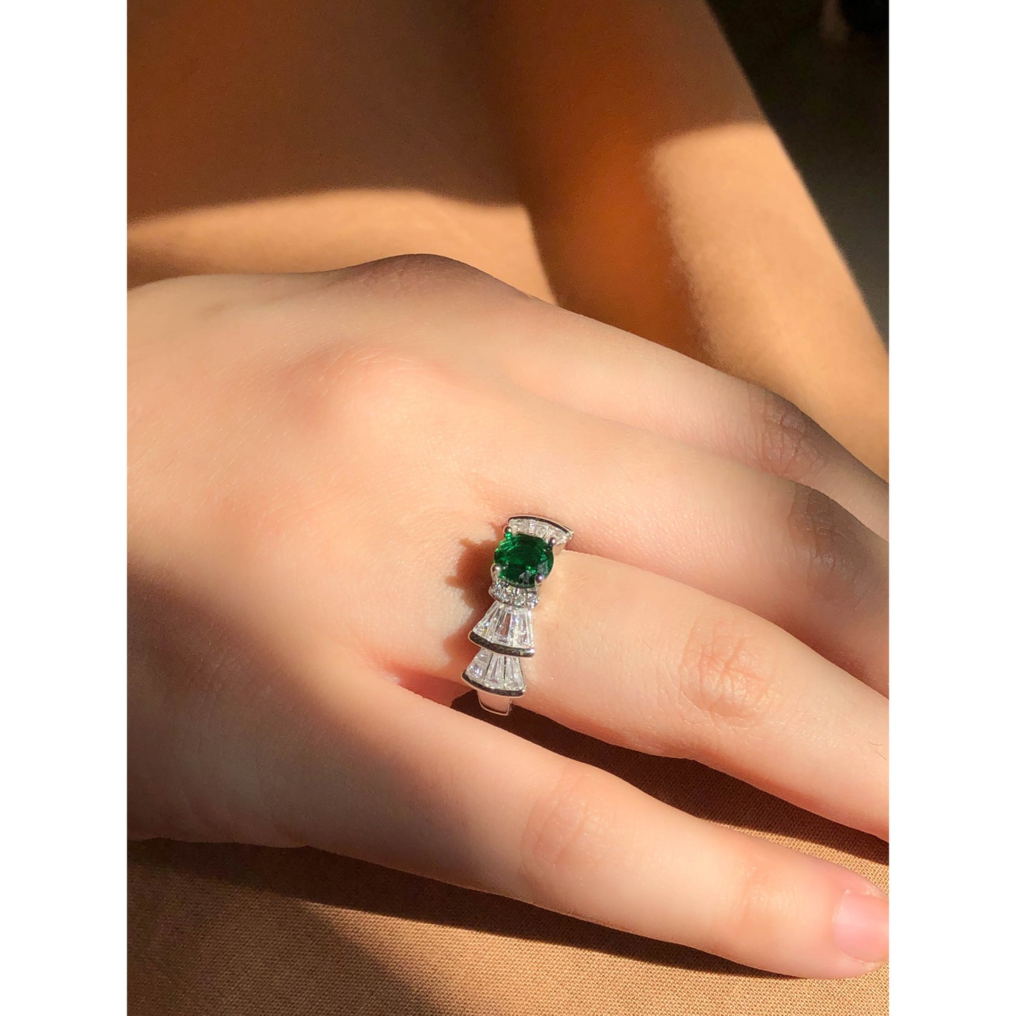 A silver-tone ring featuring an oval-cut gemstone in aqua and blush options, accented by stacked baguette-cut crystals on each side, worn on a hand with warm-toned fabric in the background.