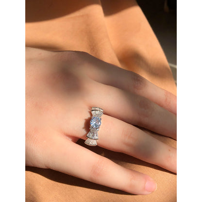 A silver-tone ring featuring an oval-cut gemstone in aqua and blush options, accented by stacked baguette-cut crystals on each side, worn on a hand with warm-toned fabric in the background.