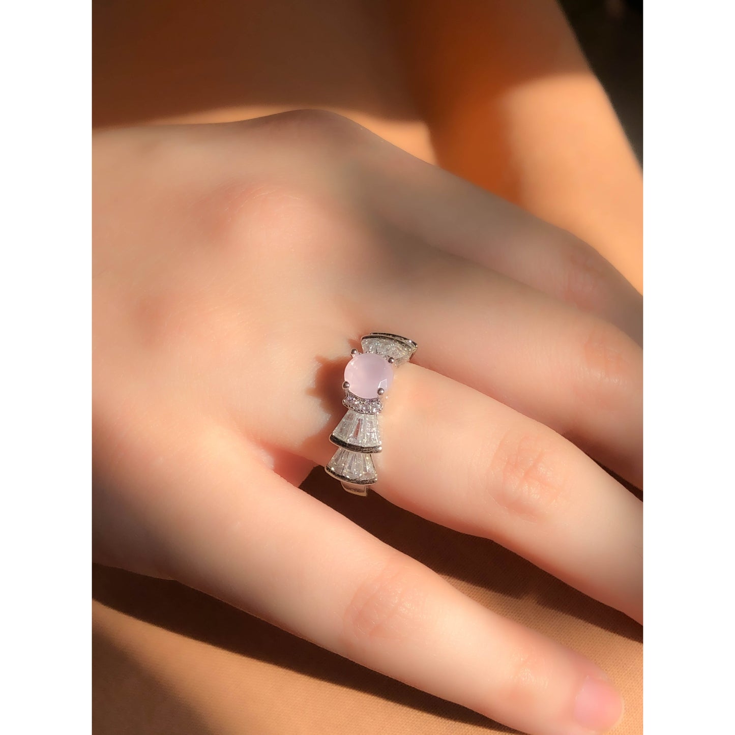 A silver-tone ring featuring an oval-cut gemstone in aqua and blush options, accented by stacked baguette-cut crystals on each side, worn on a hand with warm-toned fabric in the background.