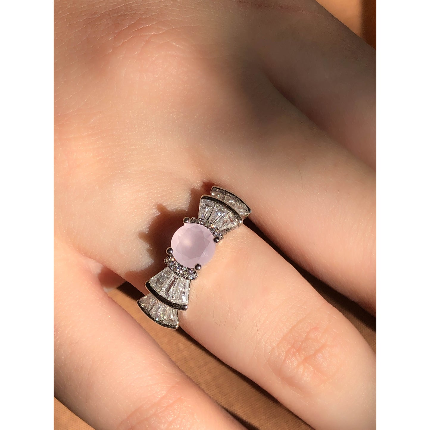 A silver-tone ring featuring an oval-cut gemstone in aqua and blush options, accented by stacked baguette-cut crystals on each side, worn on a hand with warm-toned fabric in the background.