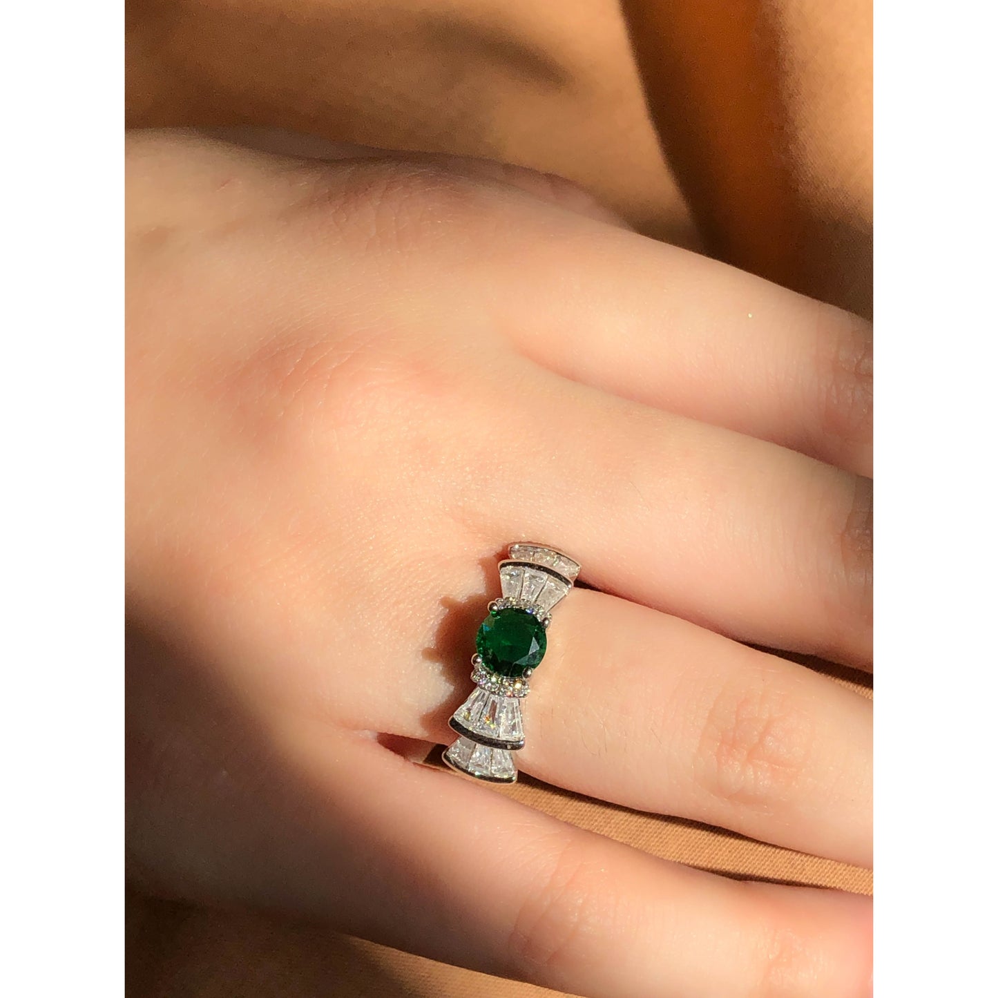 A silver-tone ring featuring an oval-cut gemstone in aqua and blush options, accented by stacked baguette-cut crystals on each side, worn on a hand with warm-toned fabric in the background.