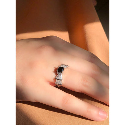 A silver-tone ring featuring an oval-cut gemstone in aqua and blush options, accented by stacked baguette-cut crystals on each side, worn on a hand with warm-toned fabric in the background.