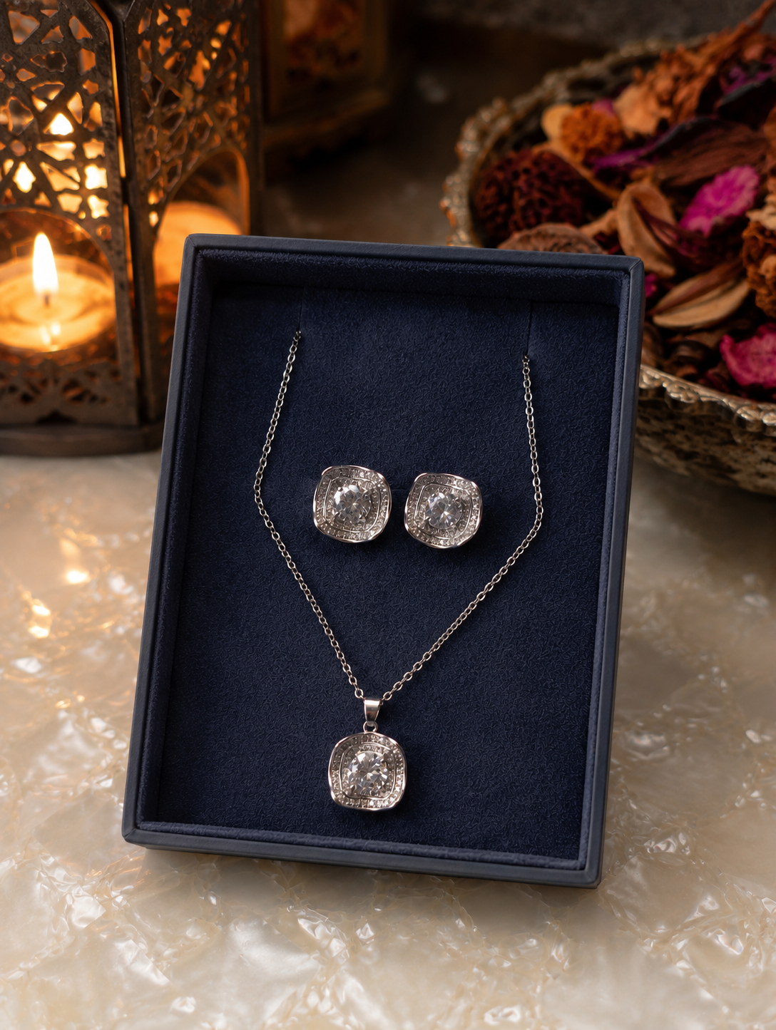 Signature silver cushion halo pendant and stud earring set displayed in a premium navy velvet jewelry box with warm candlelight and decorative floral accents