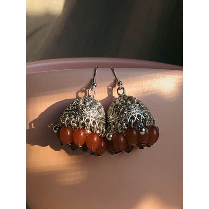 A pair of silver-tone jhumka earrings featuring a round, sparkling top section covered in clear stones, with a bell-shaped dangle below adorned with clear stones and small white pearl or bead accents