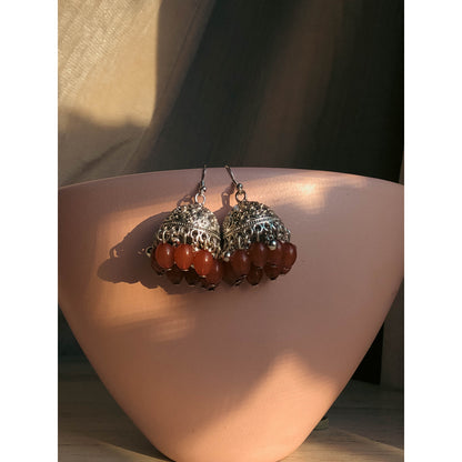 A pair of silver-tone jhumka earrings featuring a round, sparkling top section covered in clear stones, with a bell-shaped dangle below adorned with clear stones and small white pearl or bead accents