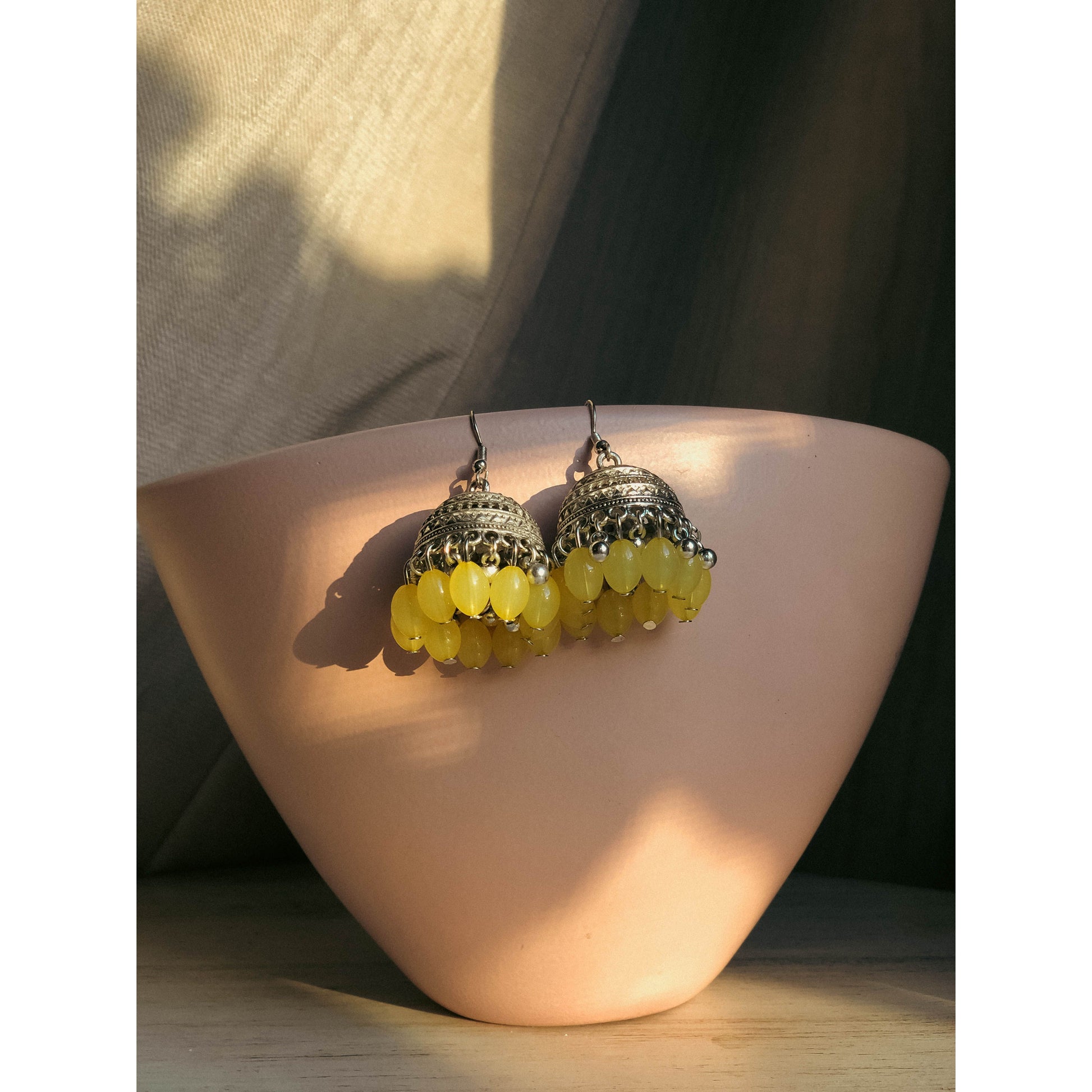 A pair of silver-tone jhumka earrings featuring a round, sparkling top section covered in clear stones, with a bell-shaped dangle below adorned with clear stones and small white pearl or bead accents