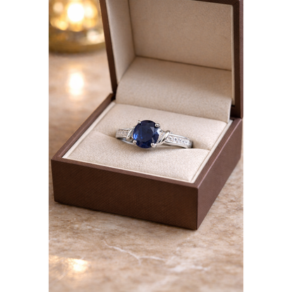 Silver ring with a blue gemstone in an open jewelry box on a marble surface.