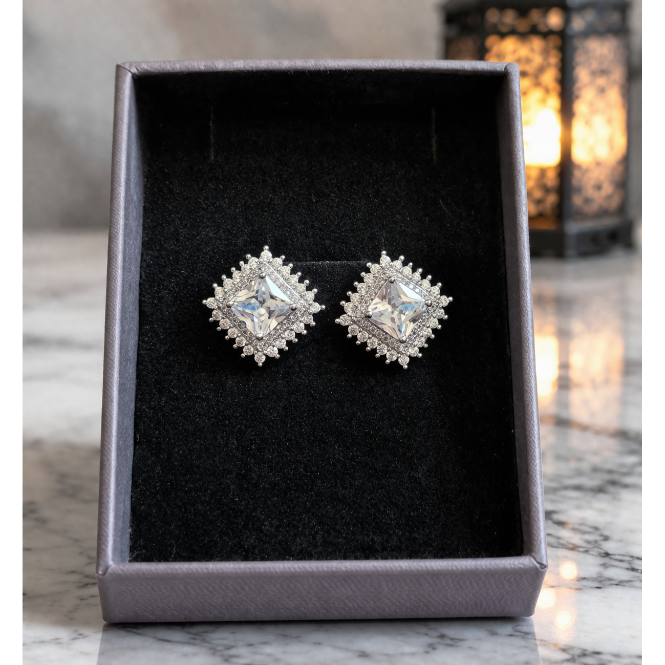 Silver square crystal stud earrings placed in grey velvet jewelry box on reflective marble surface with blurred lantern background