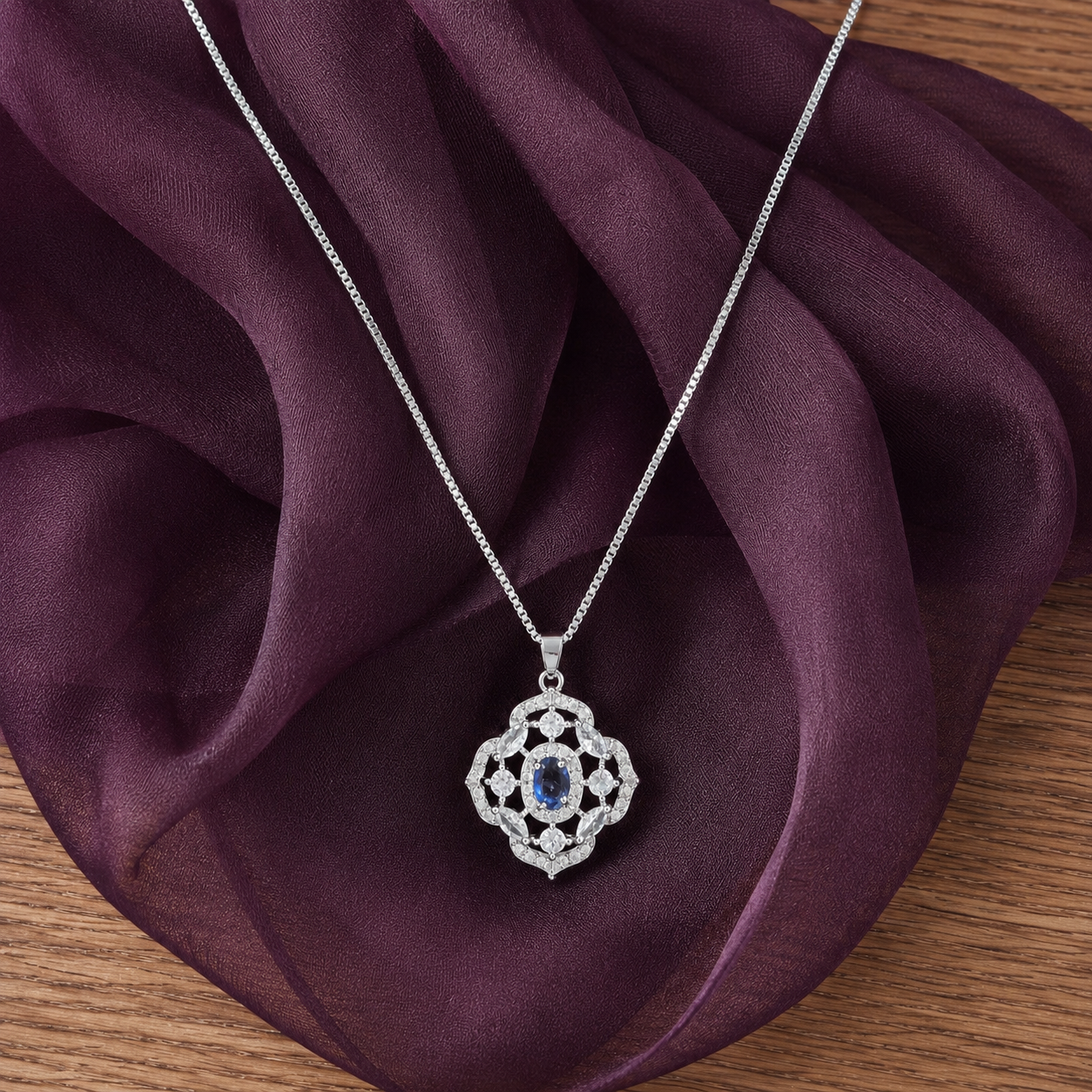 Silver-tone Nocturne Crest locket with deep blue oval centre stone and clear zircon detailing displayed on rich plum fabric.