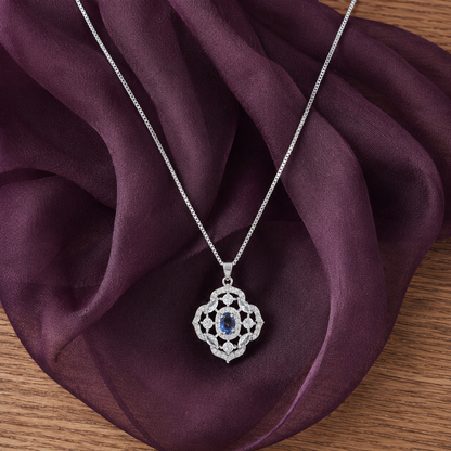Silver-tone Nocturne Crest locket with deep blue oval centre stone and clear zircon detailing displayed on rich plum fabric.