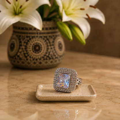 Clear stone statement ring in S925 sterling silver displayed on a beige ceramic tray with blurred lilies in the background.