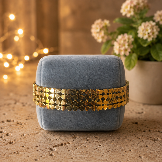 Gold floral openwork bracelet displayed around a soft smoky blue velvet bracelet cushion on a textured travertine surface with blurred indoor flowers and warm fairy lights.