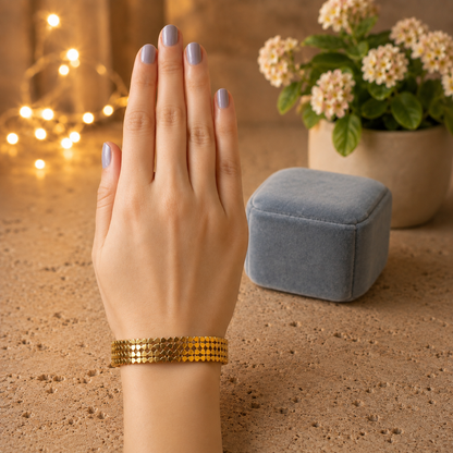 Gold floral lock bracelet worn on a hand with soft smoky blue manicure resting on a travertine surface with blurred fairy lights, flowering plant, and blue velvet bracelet cushion
