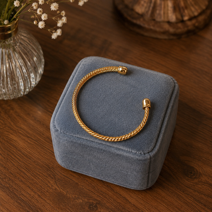 Gold open-style twisted cuff bracelet displayed on a soft blue velvet jewelry box with luxurious folds on a walnut wood surface.