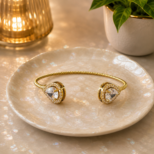 Gold open cuff bracelet with twin pear-shaped crystal stones presented on a pearlescent ceramic plate in a luxury setting.