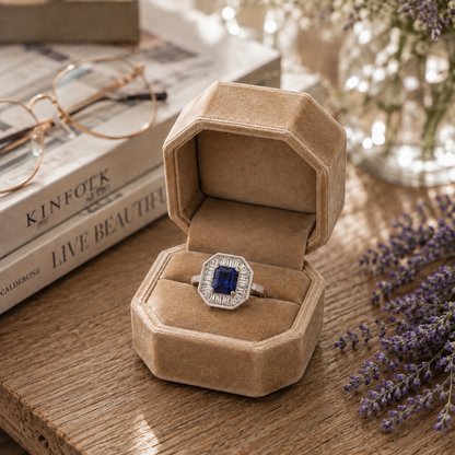 S925 sterling silver octagonal halo ring with baguette zircon accents and deep blue gem displayed inside a premium beige velvet octagonal ring box on a rustic wooden table with lavender, books, and soft ambient lighting.