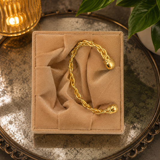 Gold braided open cuff bracelet displayed in a premium apricot velvet bracelet box on an oxidized mirror surface with warm ambient lighting.