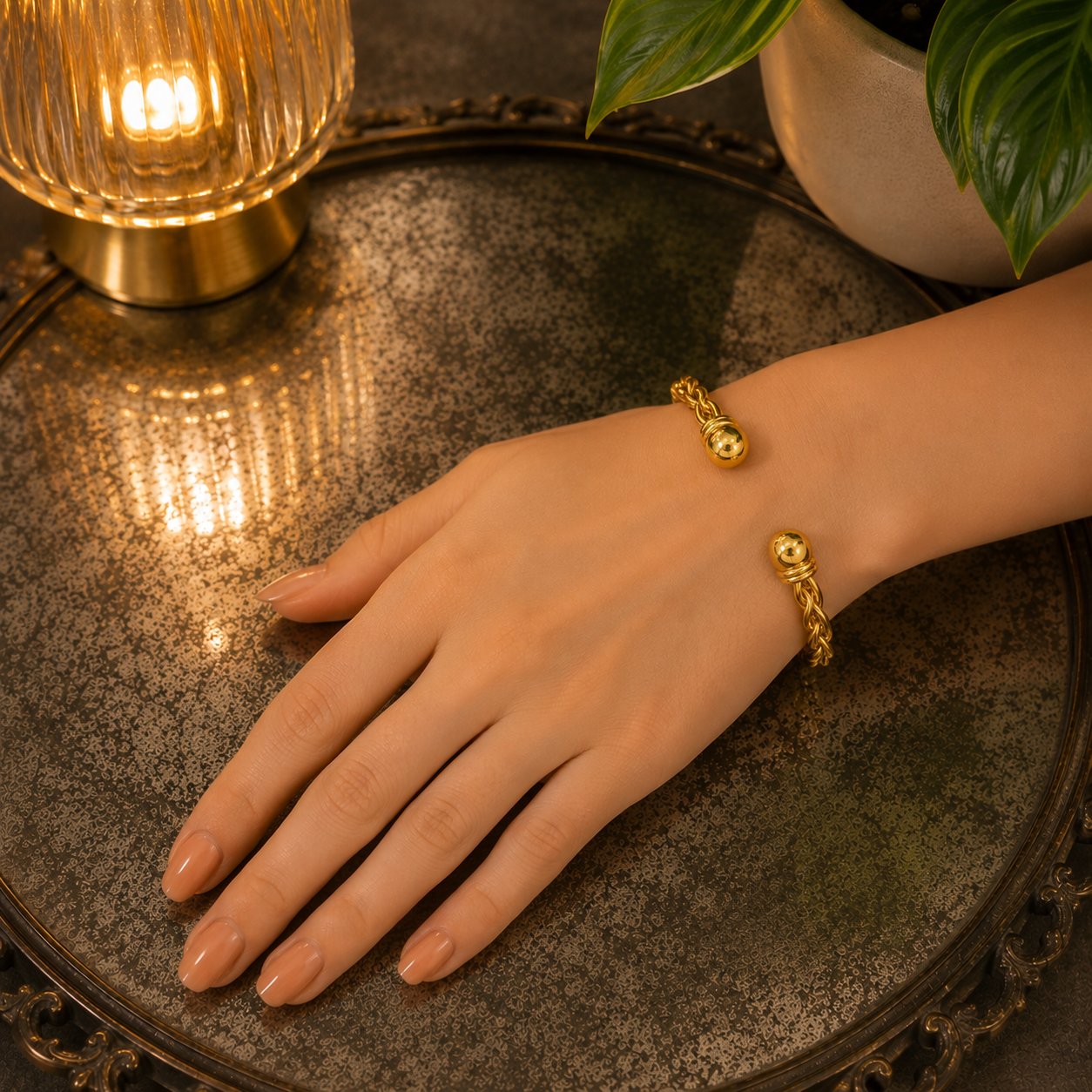 Gold braided open-style bracelet worn on a woman's wrist resting on an oxidized mirror tray with warm lamp lighting and green foliage in the background