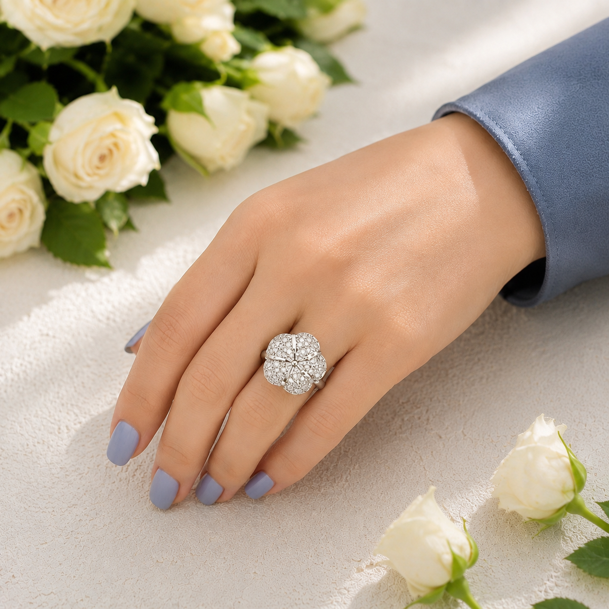 Floral crystal statement ring worn on a hand with soft blue manicure and white rose background.