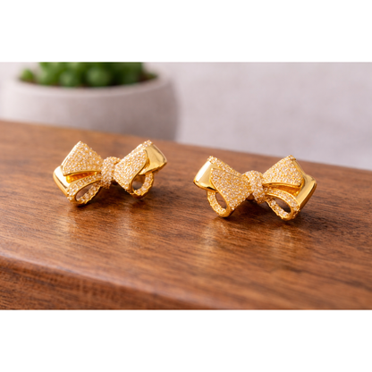 Gold bow stud earrings placed on a wooden surface with a blurred ceramic pot plant in the background, HD product shot.