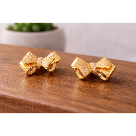Gold bow stud earrings placed on a wooden surface with a blurred ceramic pot plant in the background, HD product shot.