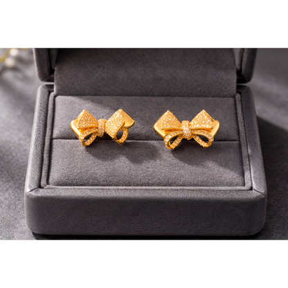 Gold bow stud earrings displayed inside a grey jewelry box with micro-pavé detailing, high-definition close-up.