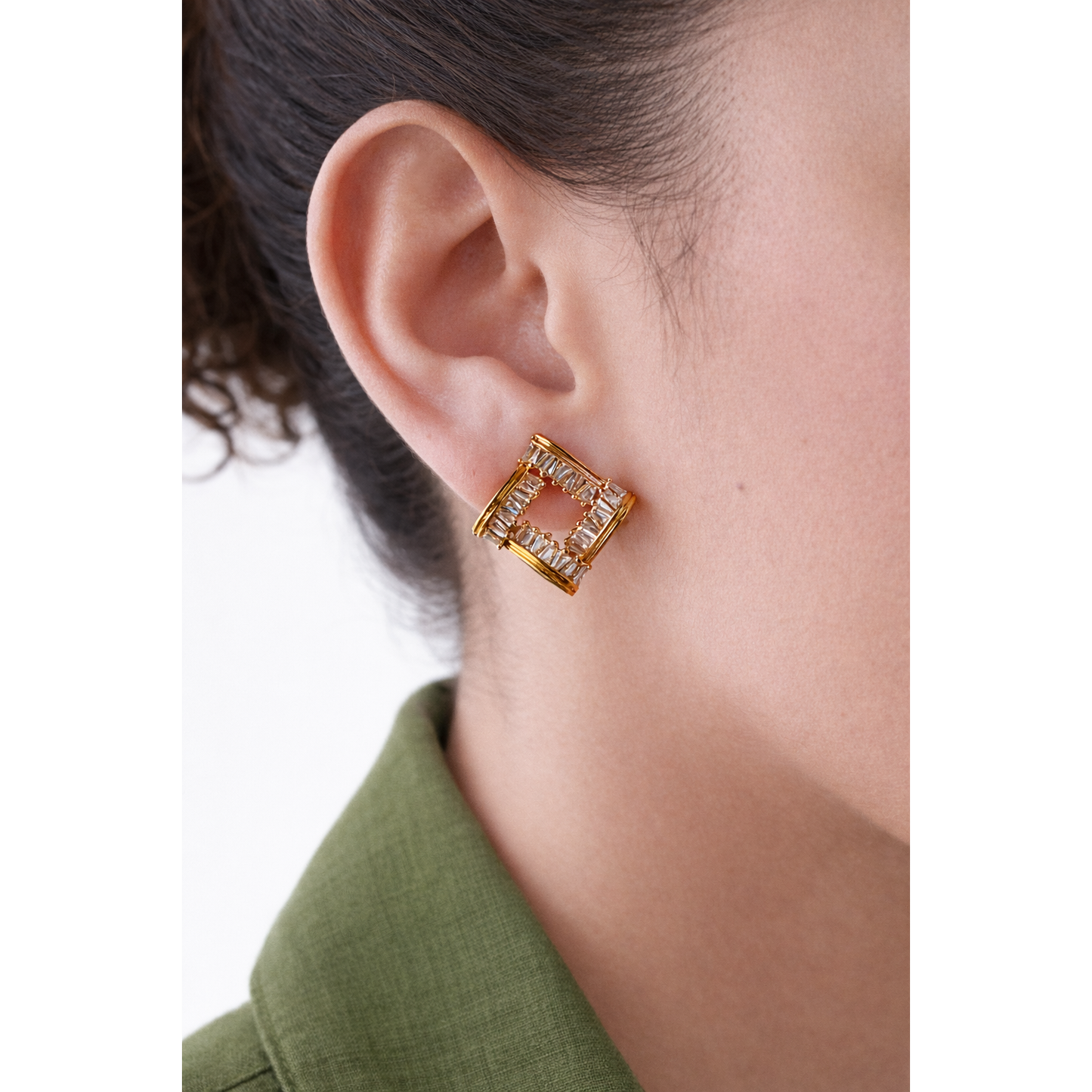 Square gold-tone baguette crystal stud earring worn on model ear with olive green linen shirt, close-up side profile jewellery photo