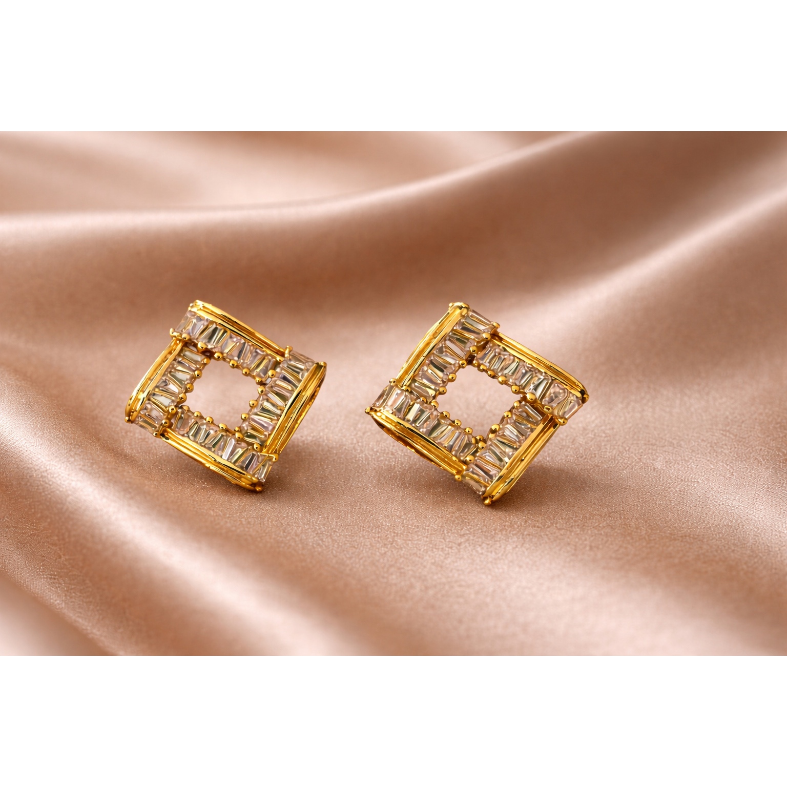 Pair of square frame gold-tone crystal stud earrings displayed on light brown satin fabric with soft folds, close-up jewellery product image