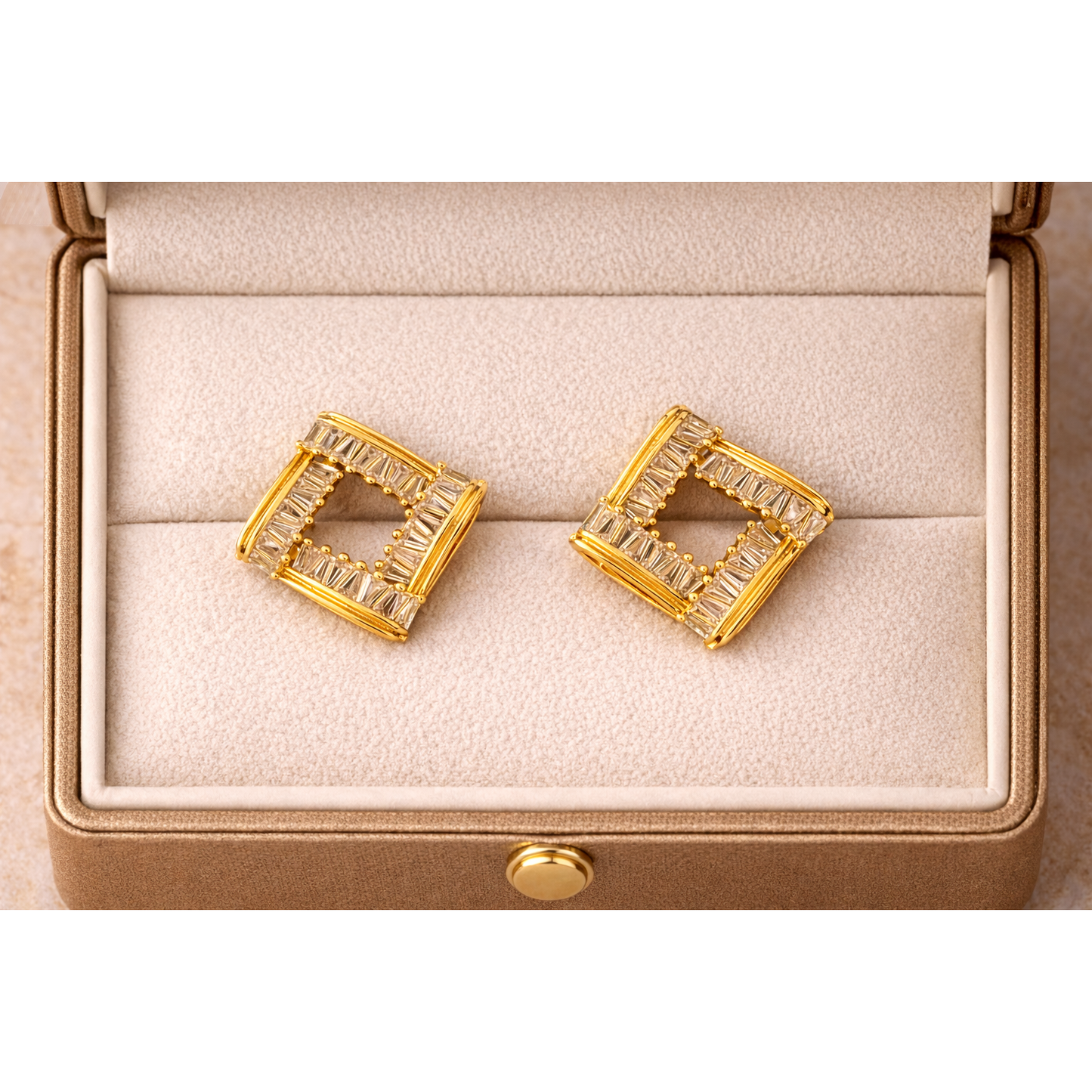 Square gold-tone baguette crystal stud earrings placed flat inside a fine jewellery box with soft cream interior, high-definition product photography