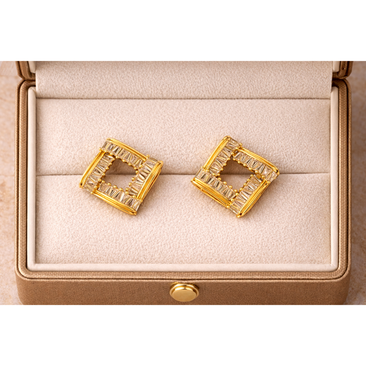 Square gold-tone baguette crystal stud earrings placed flat inside a fine jewellery box with soft cream interior, high-definition product photography